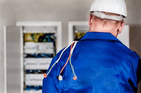 An electrician with a hard hat on with his back to the camera looking into an electrical wiring box